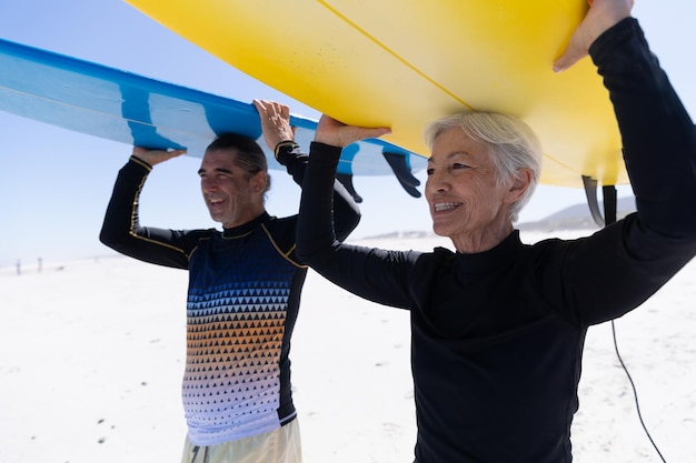 Photo senior caucasian couple enjoying time at the beach, walking towards the sea and holding surfboards above their heads