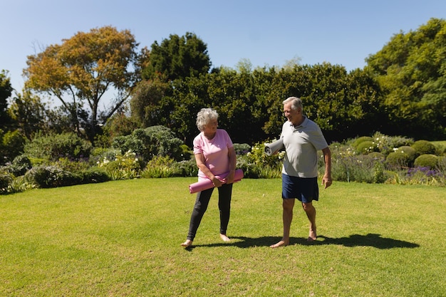 ヨガマットを持ち、日当たりの良い庭で微笑む年配の白人のカップル。退職後の隠れ家とアクティブなシニアのライフスタイルコンセプト。