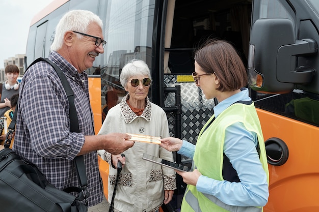 Senior caucasian couple standing at open bus door and showing tickets to bus supervisor with tablet