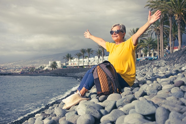 Senior caucasian gray haired woman sitting with open arms on the pebbles beach at sea looking at horizon over water Concept of vacation freedom and happy retirement