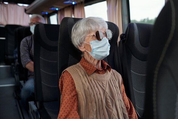 Senior caucasian lady with gray hair wearing sunglasses and facial mask sitting in bus