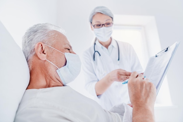 Senior caucasian man sitting on hospital bed ang signing medical form to doctor in medical masks