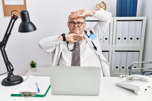 Senior caucasian man wearing doctor uniform and stethoscope at the clinic smiling cheerful playing peek a boo with hands showing face. surprised and exited