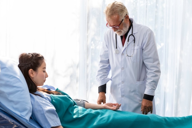 Photo senior caucasian professional doctor man check blood pressure with asian woman patient in the hospital room.