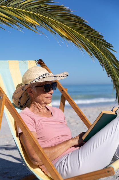 Старшая кавказская женщина наслаждается временем на пляже, сидит на шезлонге и читает книгу
