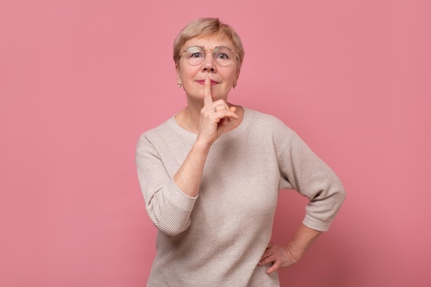 La donna caucasica anziana tiene il dito sulle labbra chiedi di stare in silenzio, guarda la fotocamera, racconta il mistero della condivisione