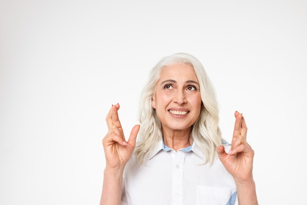 Senior caucasian woman thinking about something pleasant and making wish with crossed fingers, isolated over white wall