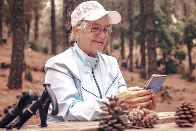 Senior caucasian woman in a trekking day in the woods sitting at a wooden table to rest using phone