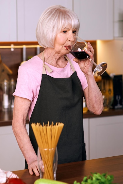 수석 쾌활한 여자는 현대 부엌에서 요리하는 동안 적포도주를 마시고 있습니다
