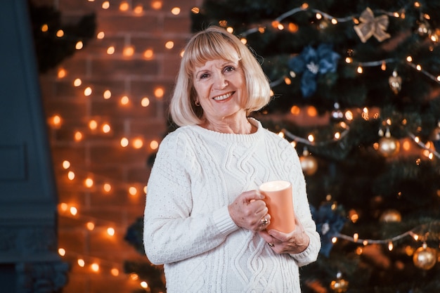 Senior cheerful woman standing with cup of drink in the room with christmas decorations.