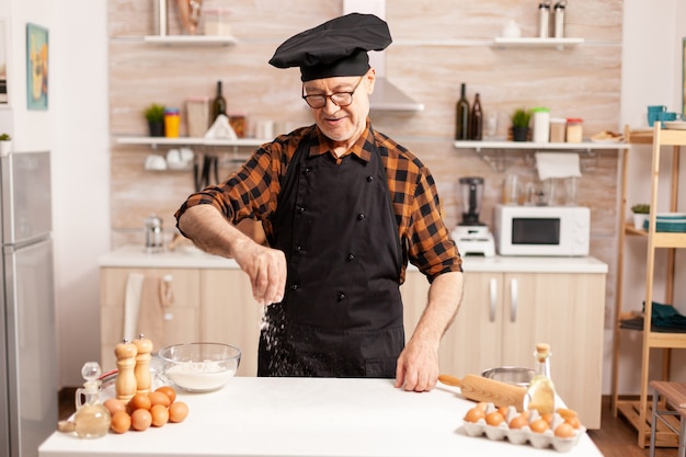 Senior chef bereidt zelfgebakken brood dat tarwemeel op de keukentafel besprenkelt. Gepensioneerde senior chef-kok met bonete en schort, in keukenuniform beregening zeven zeven ingrediënten met de hand.