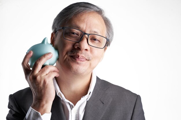 Senior chinese man with formal wear eyeglasses holding piggy bank against white background