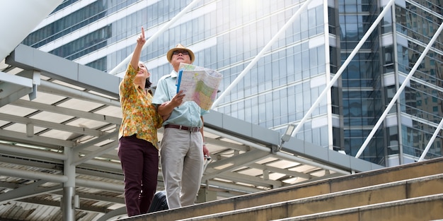 Senior couple are walking, dragging their luggage and holding a map to navigate the streets.