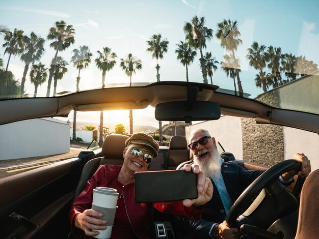 Senior couple having fun doing selfie inside a convertible car during holiday vacation Elderly travel concept