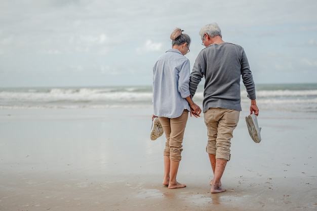 写真 手をつないでビーチに立っている年配のカップル、2人のもう一方の手は靴を持っていました。退職後のコンセプトで生命保険を計画します。