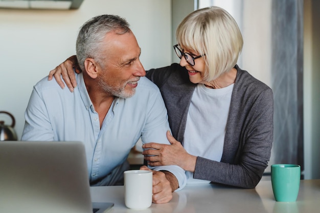 Senior couple hugging and using laptop in kitchen at home