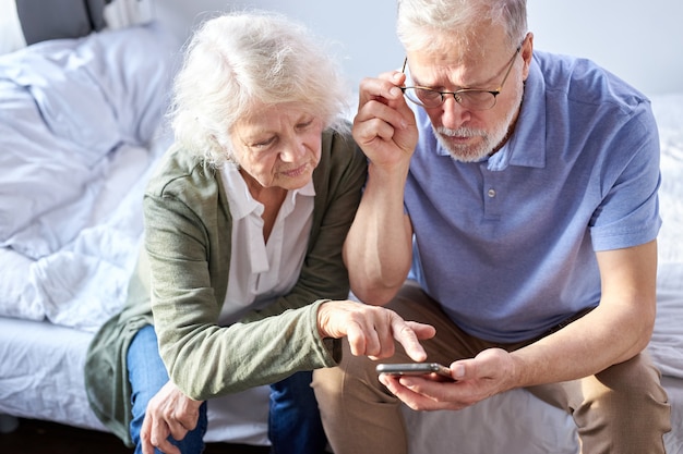 senior couple looking at children's photos in smartphone, online surfung Net,modern technology concept. caucasian woman and man using mobile phone share social media together in wellbeing home.