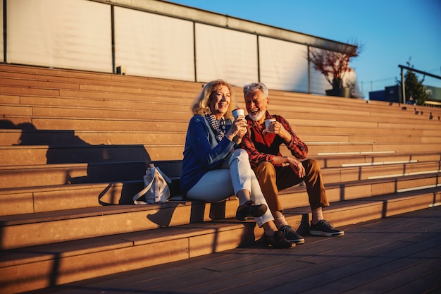 Senior couple in love sitting outdoors on the stairs and drinking coffee to go.