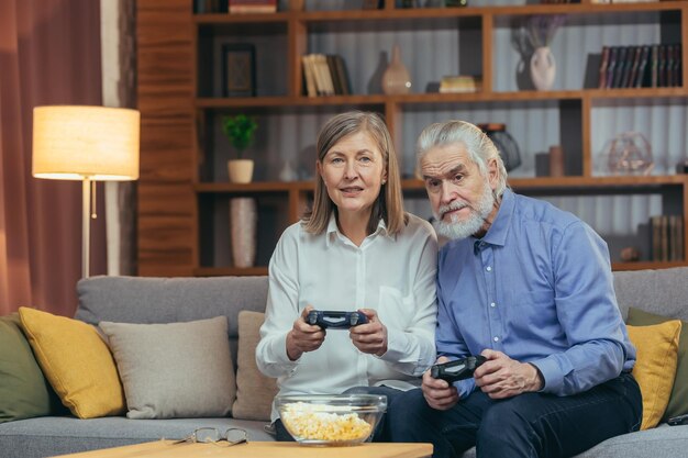 Пожилая пара играет в игровую приставку с джойстиками, сидя на диване в гостиной Смешные зрелые люди семья весело проводят свободное время вместе седые волосы мужчина и женщина наслаждаются развлечениями