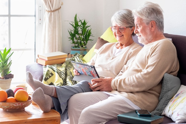 Старшие пары отдыхают на диване у себя дома, вместе глядя на цифровой планшет и улыбаясь