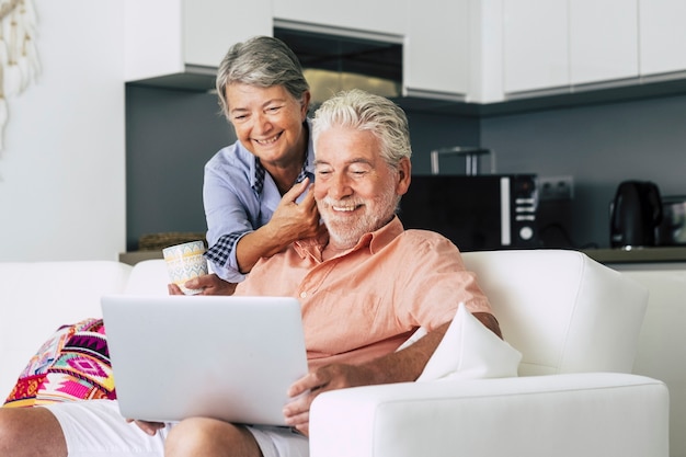 Senior couple relaxing together in the kitchen with a laptop and with a cup of coffee or tea