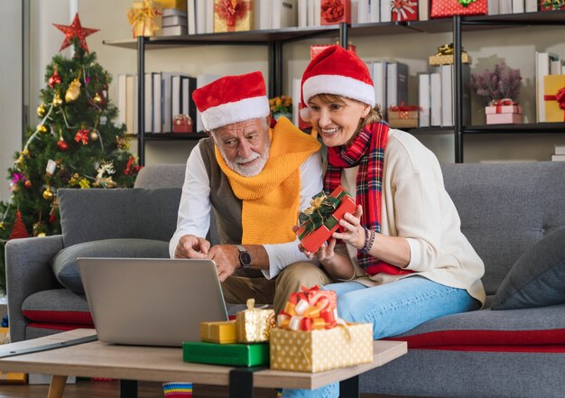 Senior couple in santa hat sitting on couch video calling\
family online using laptop computer happy with received gift at\
home with christmas tree. keeping in touch with grandchildren on\
holidays.