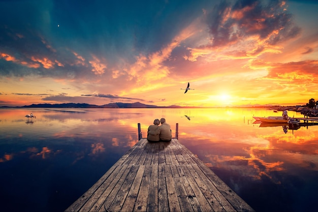 Photo senior couple seated on a wooden jetty looking a colorful sunset on the sea