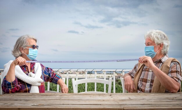 Coppia senior seduta all'aperto al tavolo di legno che indossa una maschera con in mano un metro per la distanza sociale - pensionamento di due nonni ai tempi del coronavirus, orizzonte sull'acqua sullo sfondo