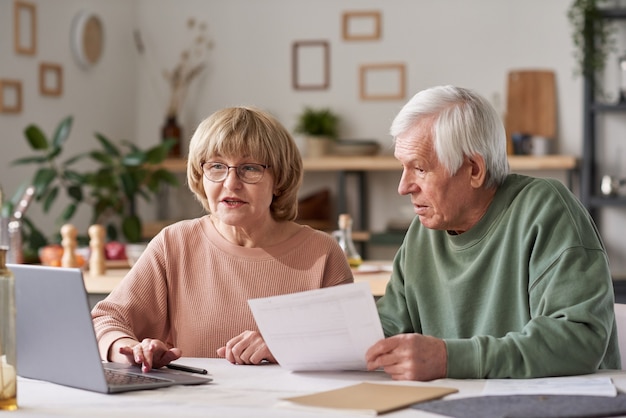 Senior couple sitting at the table and using laptop to pay for bills online in the kitchen