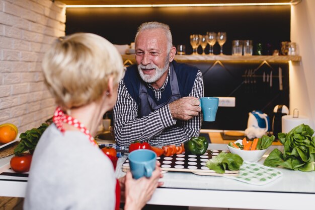 キッチンで一緒に話し、コーヒーを飲み、昼食を準備する年配のカップル。