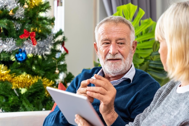 Coppia senior utilizzando tablet per videochiamare per salutare la famiglia per il festival di natale seduto sul divano con decorazioni e albero