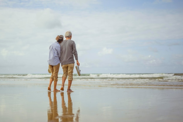 日の出で手をつないでビーチを歩いている年配のカップルは、退職の概念で生命保険を計画します。