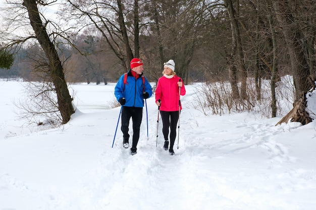 雪の降る冬の森でノルディックウォーキングポールを持って歩く年配のカップル