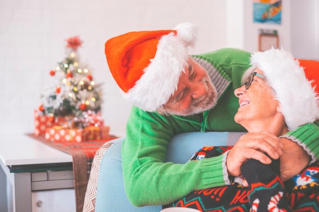 自宅で飾られたクリスマスツリーの前でお互いの手を握って暖かい服とサンタの帽子をかぶった年配のカップル。一緒にクリスマスフェスティバルを祝う愛する古いロマンチックな異性愛者のカップル