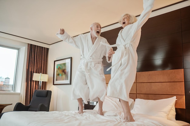 Senior couple in white robes jumping on bed and looking happy