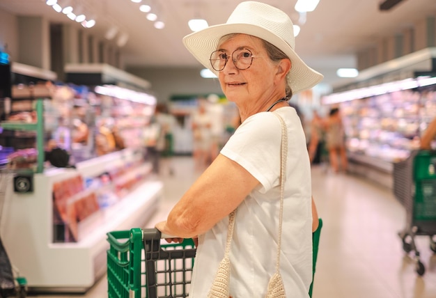 senior customer woman with hat pushing cart in supermarket pays attention to increase in prices