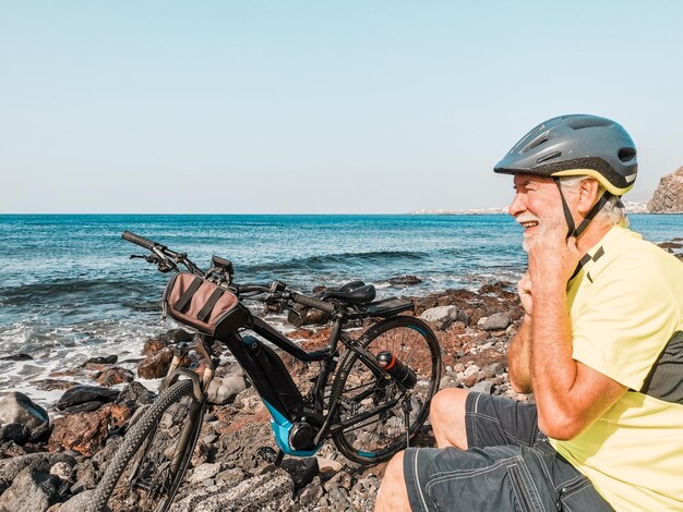 야외에서 자전거 타는 노인은 바다, 자유, 건강한 생활 방식을 즐기는 헬멧을 닫고 타는 것을 멈춥니다.