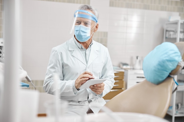 Senior die een laboratoriumjas en een beschermend masker draagt met een tablet terwijl hij in de tandheelkundige kliniek werkt