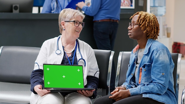 Senior doctor showing laptop with greenscreen template to\
patient sitting in waiting area lobby. medic and person looking at\
blank copyspace with chroma key display and isolated mockup.