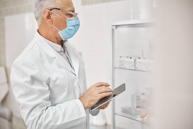 Medico anziano che indossa una maschera e tiene in mano un tablet mentre guarda un armadietto di vetro pieno di medicine