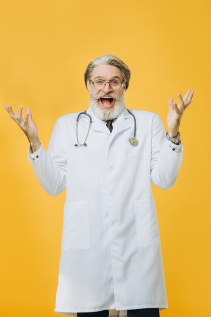 Senior doctor wearing white coat and stethoscope isolated on yellow wall raised his hands up in surprise and happiness