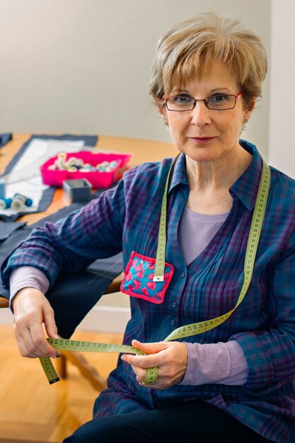 Senior dressmaker posing looking at camera with table with sewing materials in the background