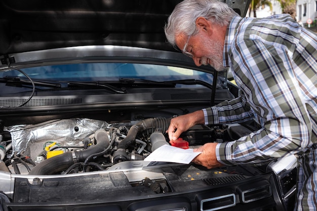 Senior driver man stopped in the street for a car breakdown with the hood open looks at the instruction booklet to understand what is broken