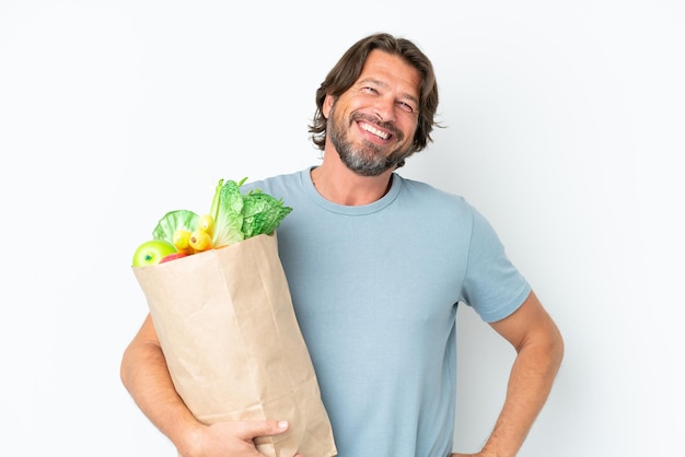 Senior dutch man holding grocery shopping bag over isolated background posing with arms at hip and smiling