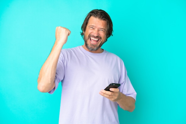Photo senior dutch man isolated on blue background with phone in victory position