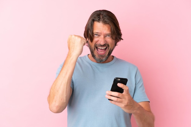 Senior dutch man isolated on pink background using mobile phone and doing victory gesture