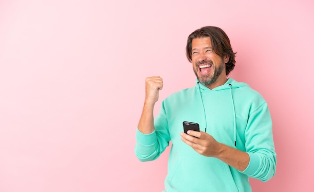 Senior dutch man isolated on pink background with phone in victory position