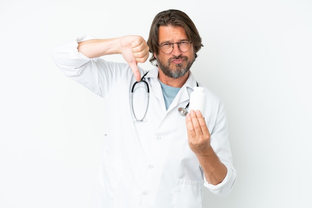 Senior dutch man isolated on white background wearing a doctor gown and holding pills while doing bad signal