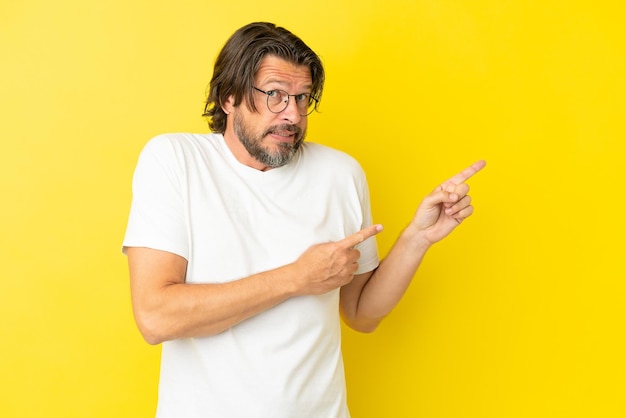 Senior dutch man isolated on yellow background frightened and pointing to the side