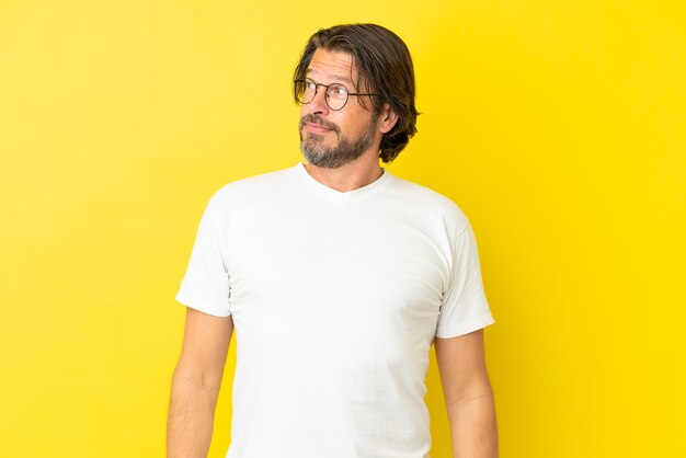 Senior dutch man isolated on yellow background making doubts gesture looking side